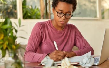 woman writing on sheet of paper with computer